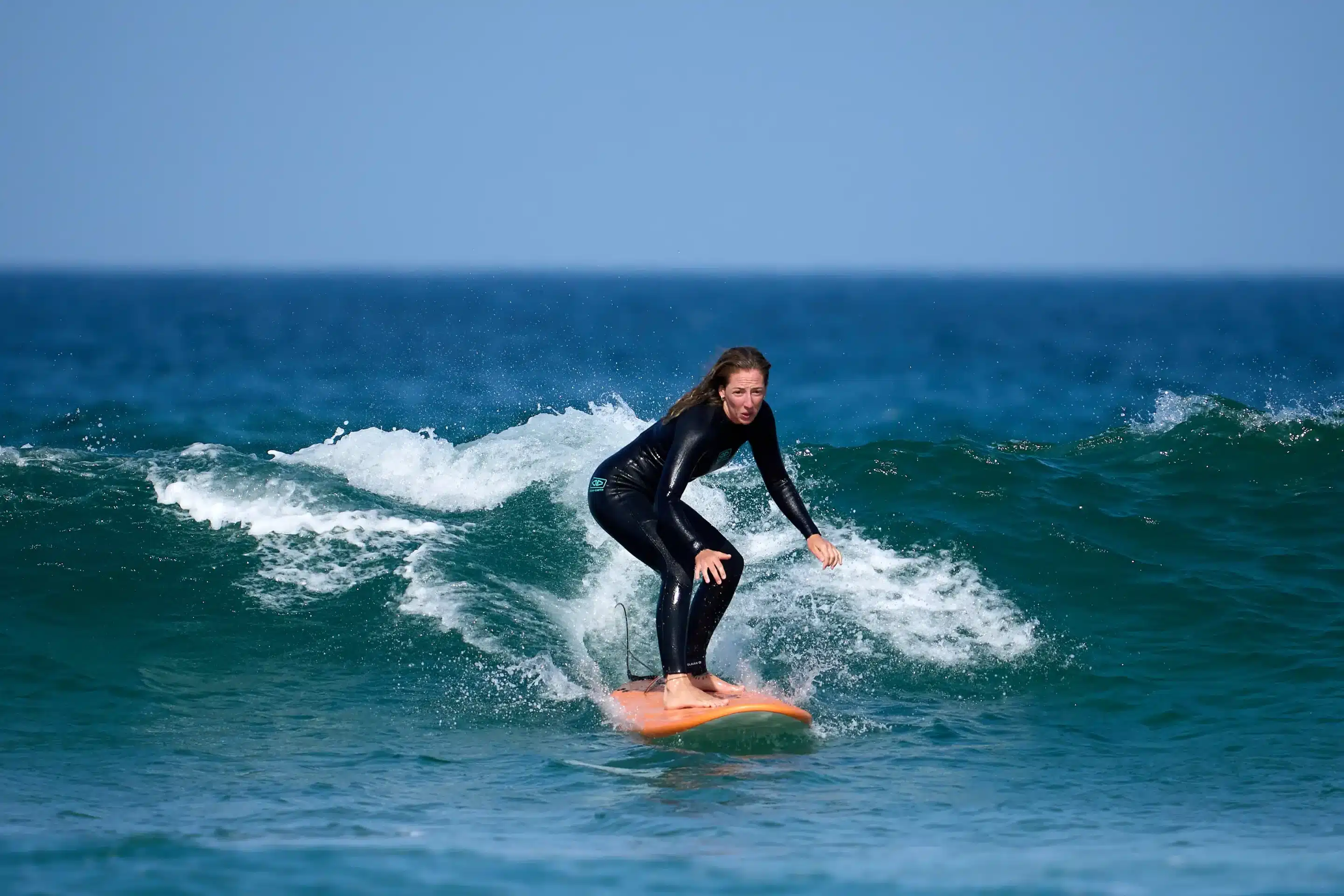 Student surfing a wave during a surf lesson at Costa da Caparica, wearing a wetsuit and riding an orange foam board — Gecko Surf School.