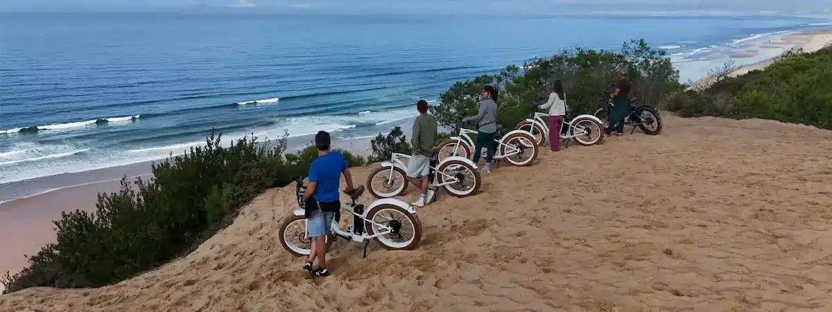 Group on an e-bike tour on the dunes of Costa da Caparica overlooking the Atlantic Ocean Group of people with white electric fat bikes on the dunes of Costa da Caparica looking out over the Atlantic Ocean on a Gecko Bike Tours guided tour