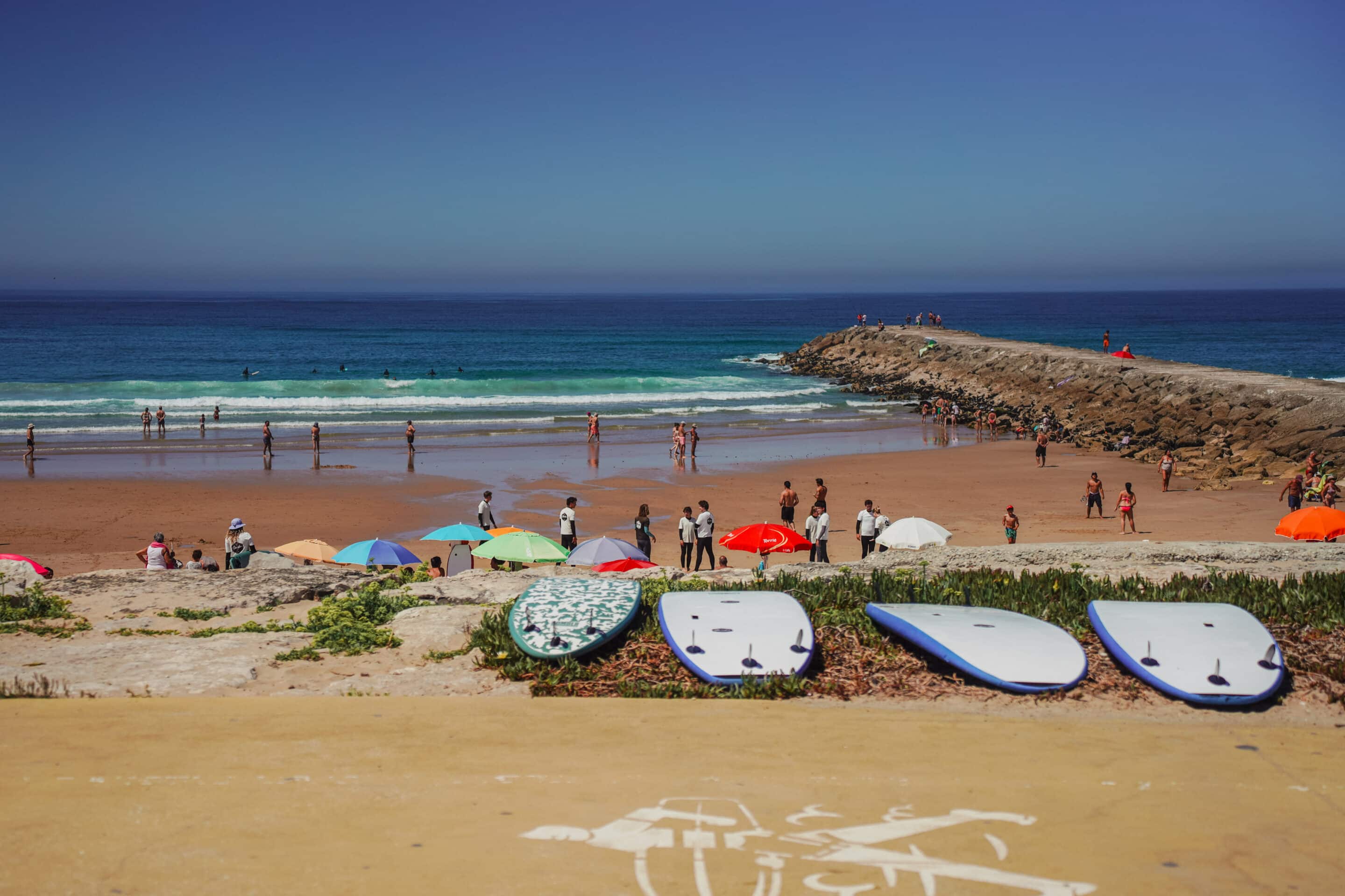 Pranchas de surf no areal da Costa da Caparica com banhistas, ondas e o molhe ao fundo num dia de verão